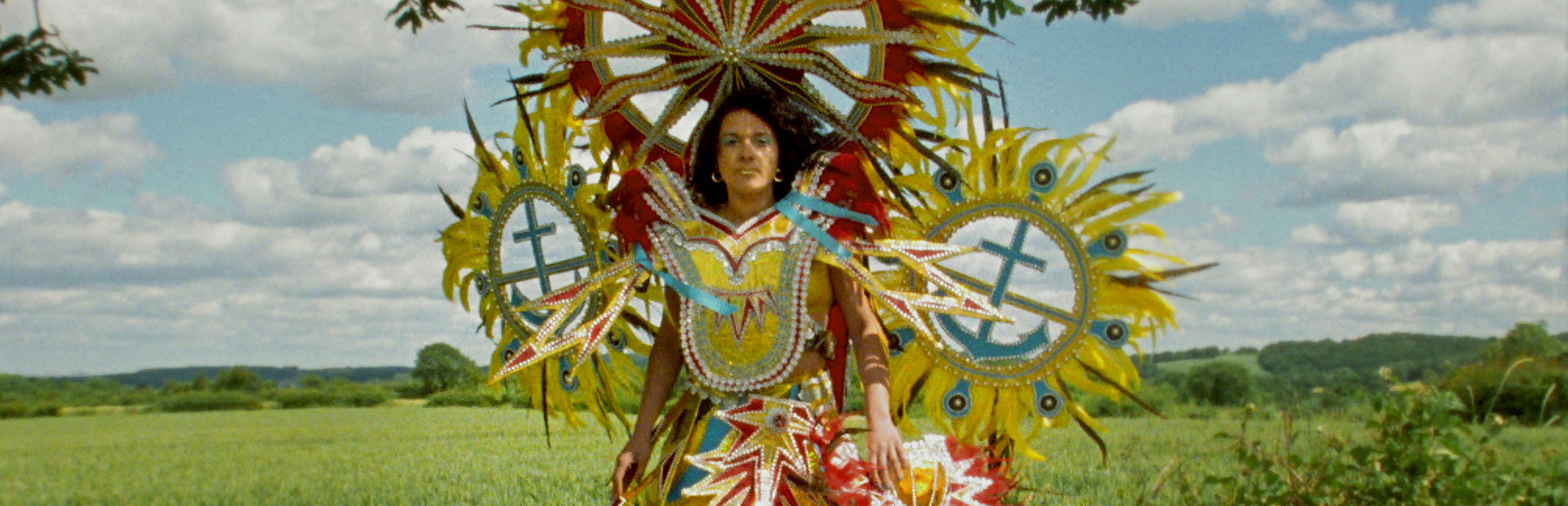 Film still of Rhea Storr's 'A Protest, A Celebration, A Mixed Message' depicting a woman wearing a large, colorful Carnival costume while standing on a wooden bench in front of a green field.