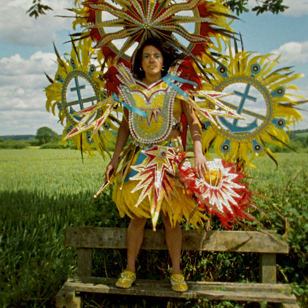 Film still of Rhea Storr's 'A Protest, A Celebration, A Mixed Message' depicting a woman wearing a large, colorful Carnival costume while standing on a wooden bench in front of a green field.