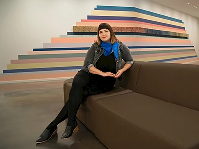 Photo of Risa Hricovsky sitting on a couch in front of a colorful art installation.