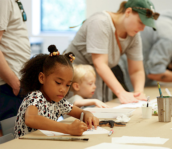 Photo of a young girl drawing in an art studio with children and adults doing the same in the background.
