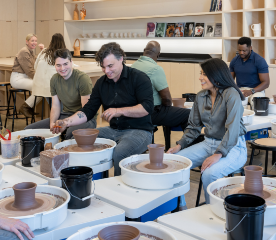 Photo of an instructor throwing clay on a potter's wheel between a man and woman in a pottery studio.