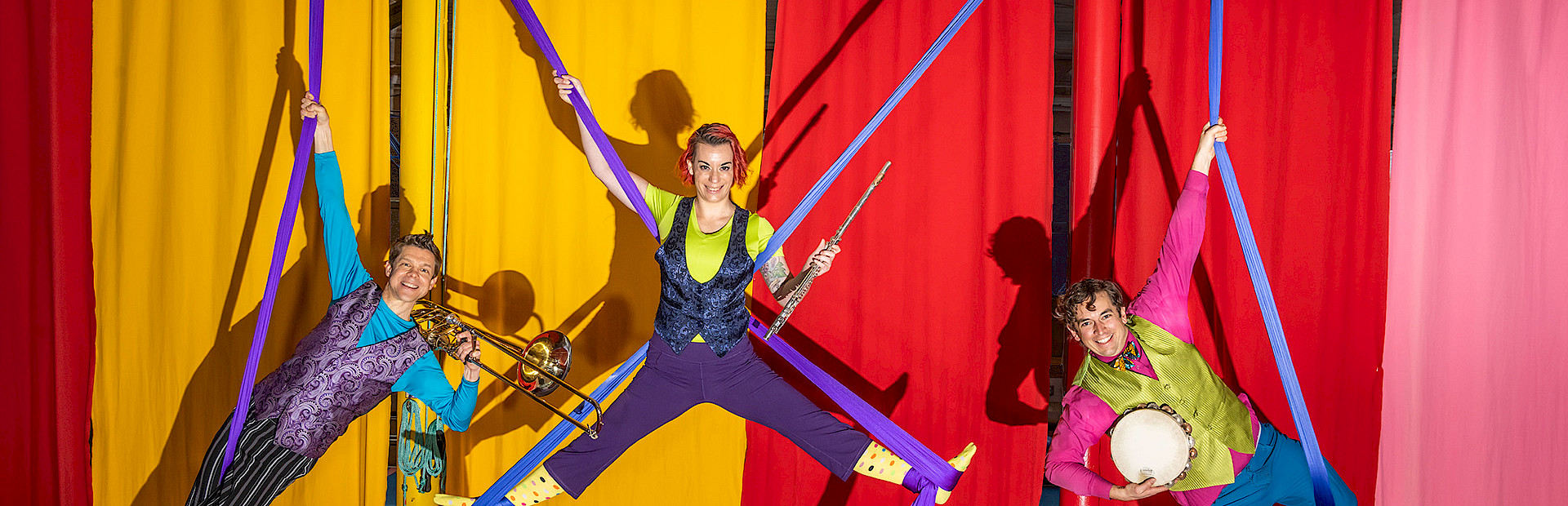 Photo of a trumpet player, flute player, and tambourine player hanging from purple silks above a stage with red and yellow curtains behind them.