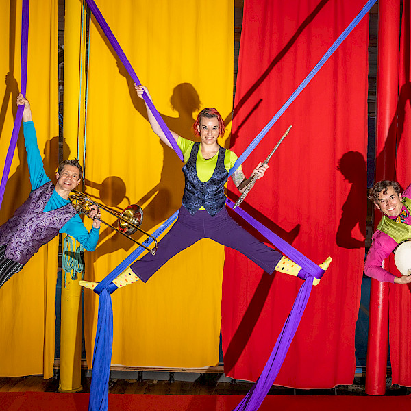 Photo of a trumpet player, flute player, and tambourine player hanging from purple silks above a stage with red and yellow curtains behind them.