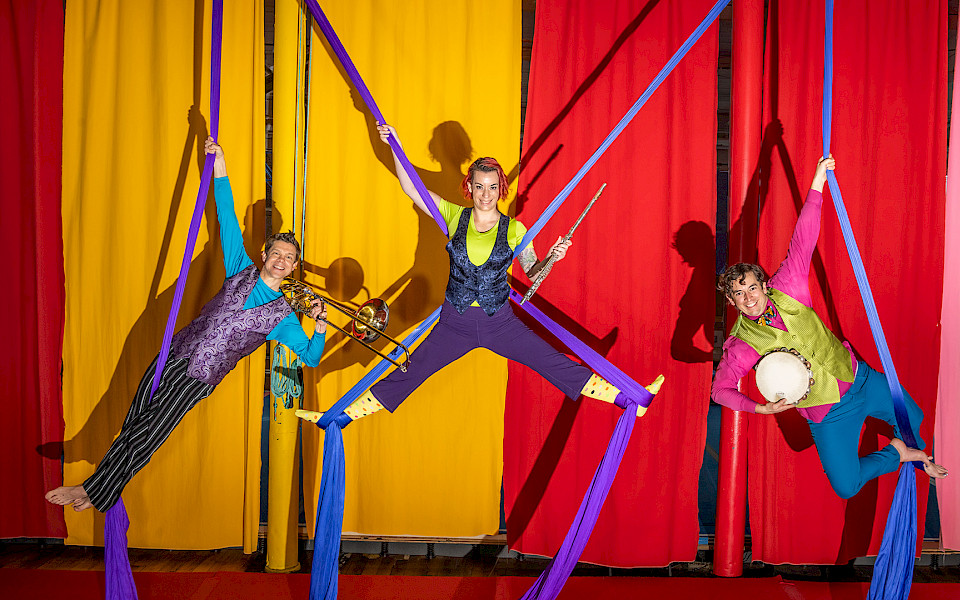 Photo of a trumpet player, flute player, and tambourine player hanging from purple silks above a stage with red and yellow curtains behind them.