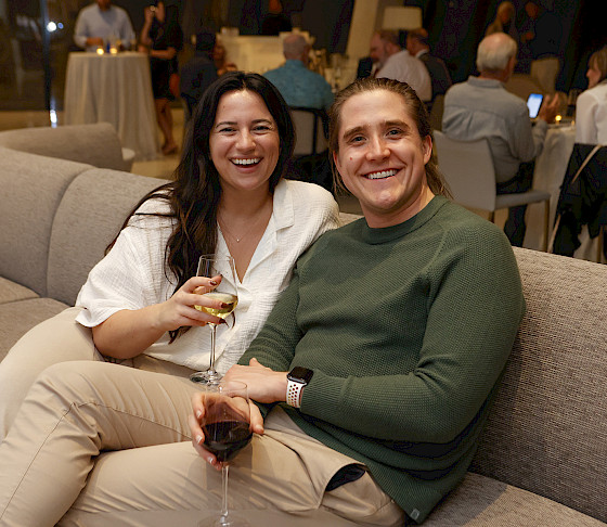 Photo of a man and woman smiling and holding wine glasses while seated on a gray couch in the Cultural Living Room.