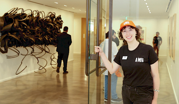 Photo of a woman wearing an AMFA T-shirt and hat holding open the door to an art gallery.