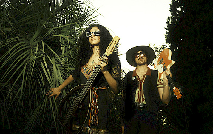 Photo of a man and woman wearing sunglasses and holding guitars while walking next to tropical trees.