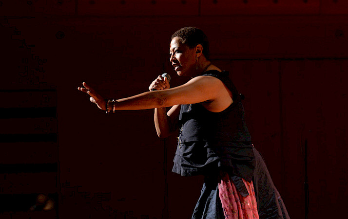 Photo of a woman on a stage singing into a microphone in her right hand and gesturing out towards the audience with her left hand.