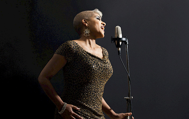 Photo of a woman with short light hair wearing a leopard print dress and dangly earrings singing into a microphone.