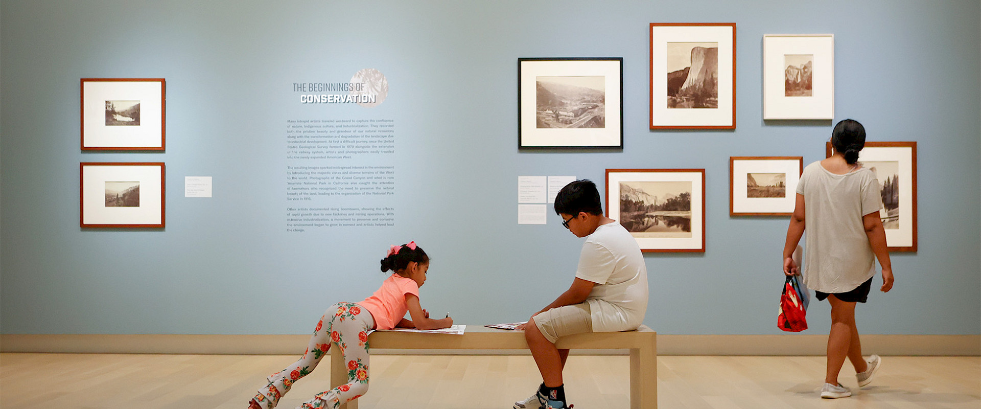 Photo of a woman looking at several framed landscape photographs on a wall in an art gallery while her two children sit on a bench nearby drawing.