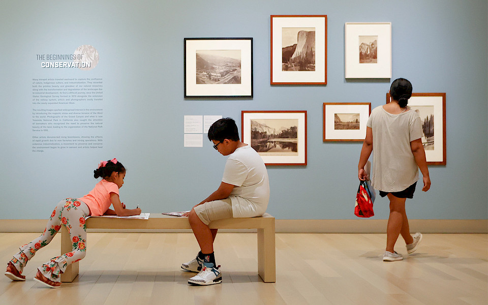 Photo of a woman looking at several framed landscape photographs on a wall in an art gallery while her two children sit on a bench nearby drawing.