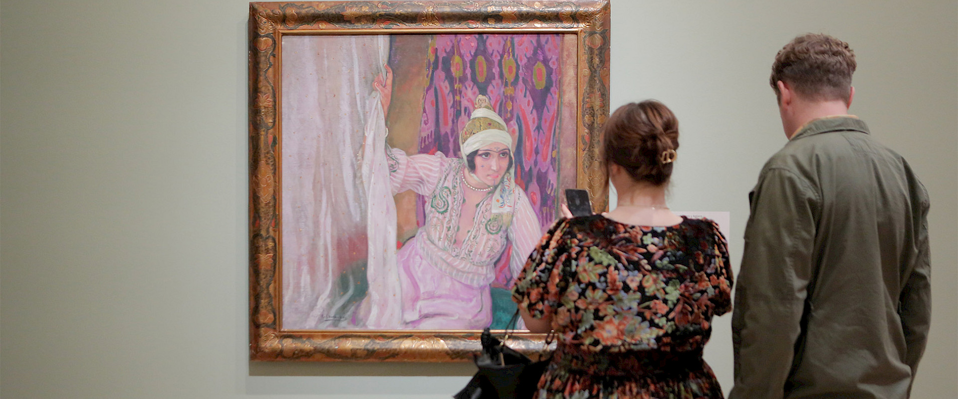 Photo of a man and a woman looking at a painting in a gilded frame on a wall in an art gallery.