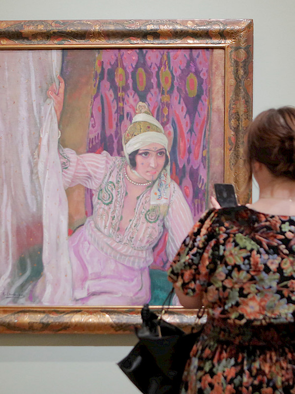 Photo of a man and a woman looking at a painting in a gilded frame on a wall in an art gallery.