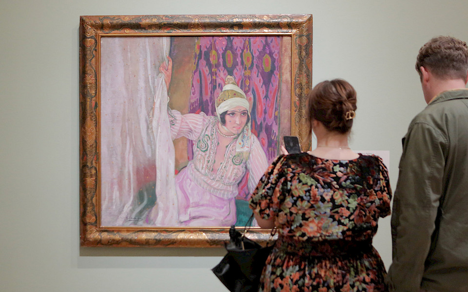 Photo of a man and a woman looking at a painting in a gilded frame on a wall in an art gallery.