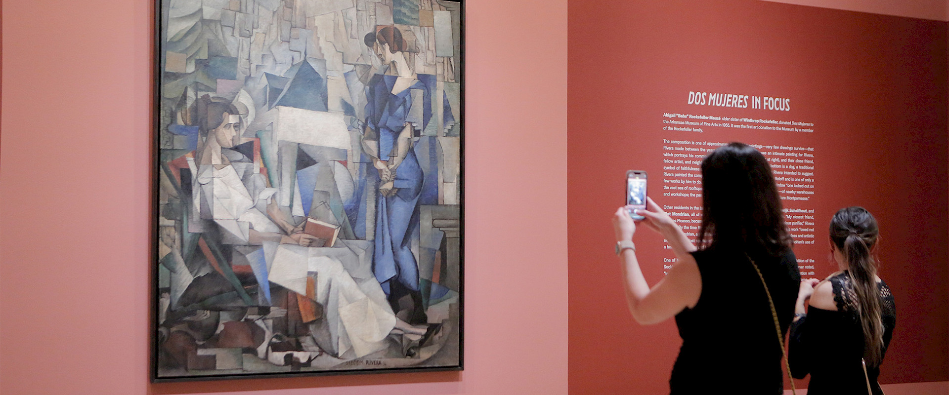Photo of two women standing in front of a painting of two women in an art gallery. The woman on the left is taking a photo of the painting with her cellphone.