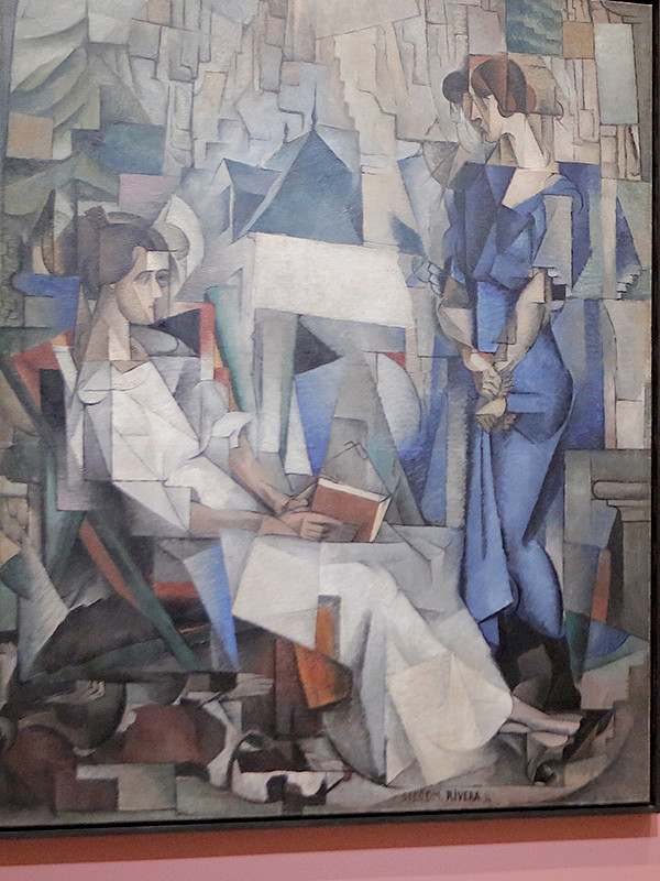 Photo of two women standing in front of a painting of two women in an art gallery. The woman on the left is taking a photo of the painting with her cellphone.