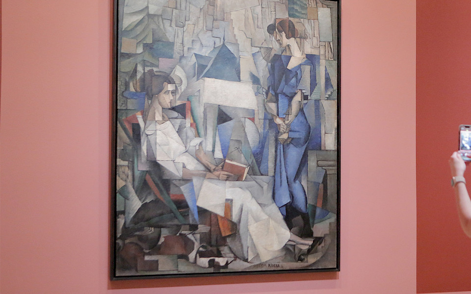 Photo of two women standing in front of a painting of two women in an art gallery. The woman on the left is taking a photo of the painting with her cellphone.