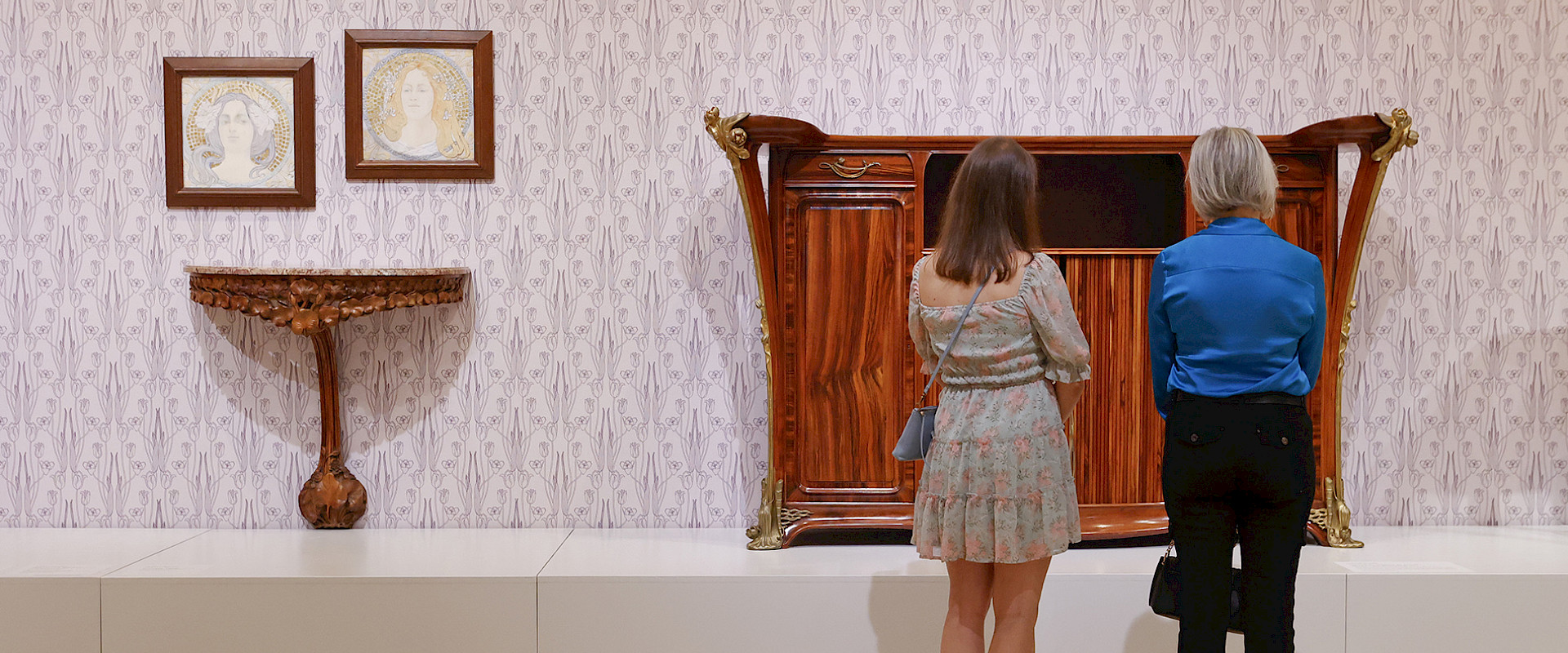 Photo of two women looking at Art Nouveau furniture in a gallery at the Arkansas Museum of Fine Arts.