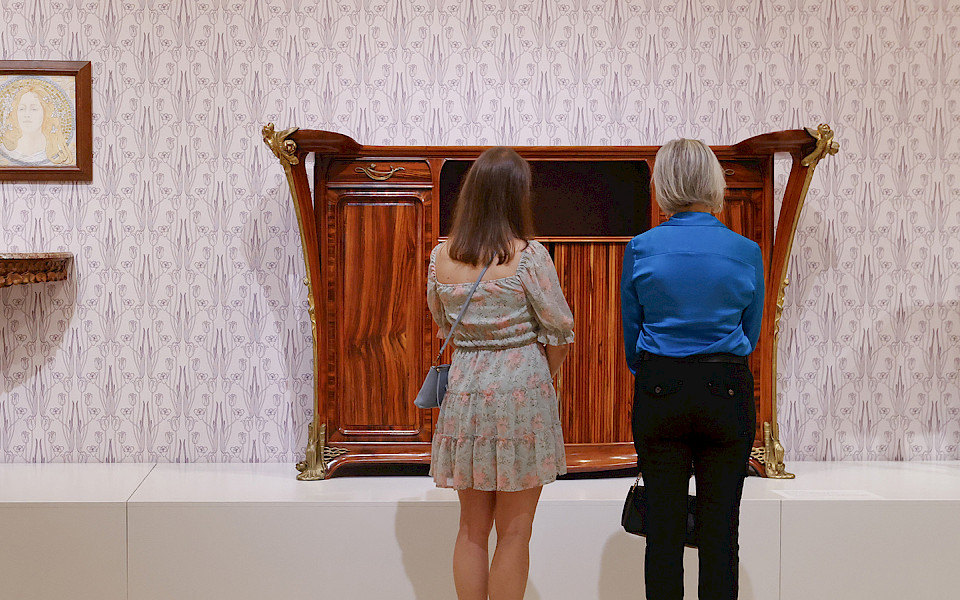 Photo of two women looking at Art Nouveau furniture in a gallery at the Arkansas Museum of Fine Arts.