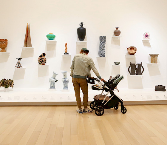 Photo of a man pushing a baby stroller while looking at sculptures on a wall in an art gallery.