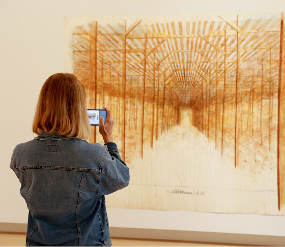 Photo of a woman taking a picture of a drawing hanging on the wall of an art gallery with her smartphone.