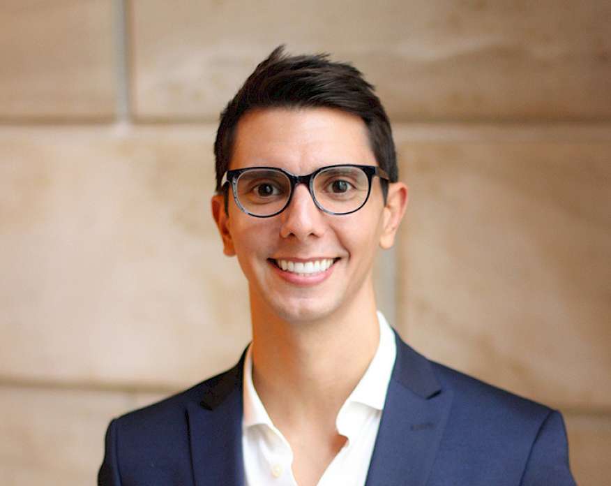 Photo of a smiling man wearing a white shirt, navy blue jacket, and glasses.