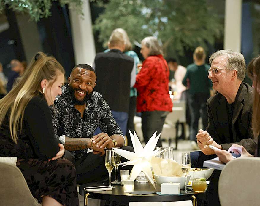 Photo of two men and two women sitting an a table drinking champagne with other people in the background.