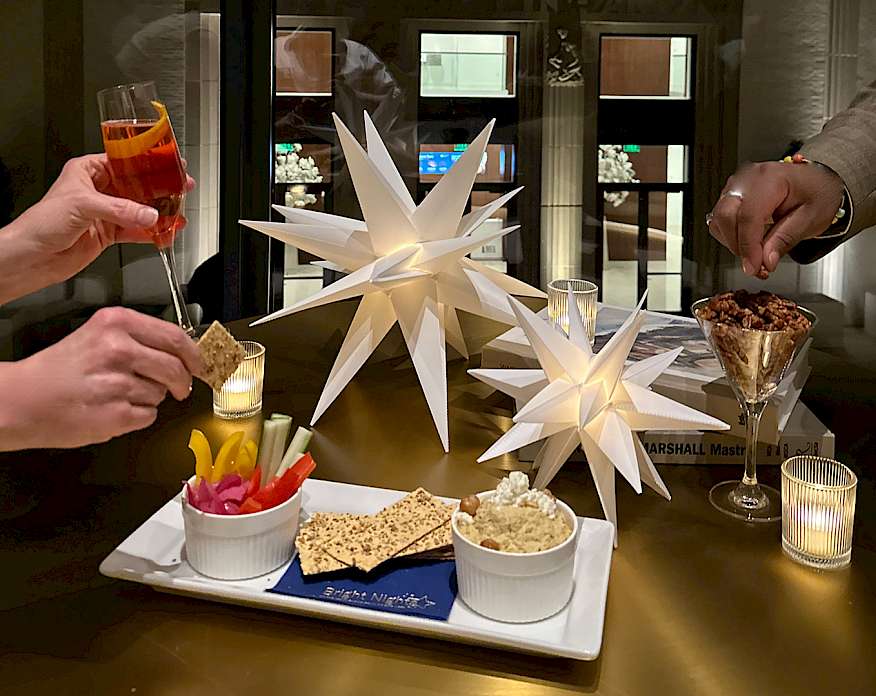 Photo of two sets of hands holding cocktails and reaching for snacks on a table decorated with votive candles and paper stars.