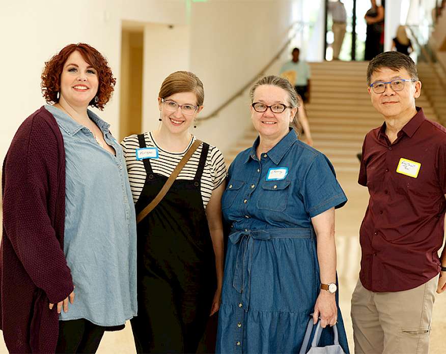 Photo of a three women and one man posed smiling together in the Atrium at AMFA. All four people are smiling and wearing name tags.