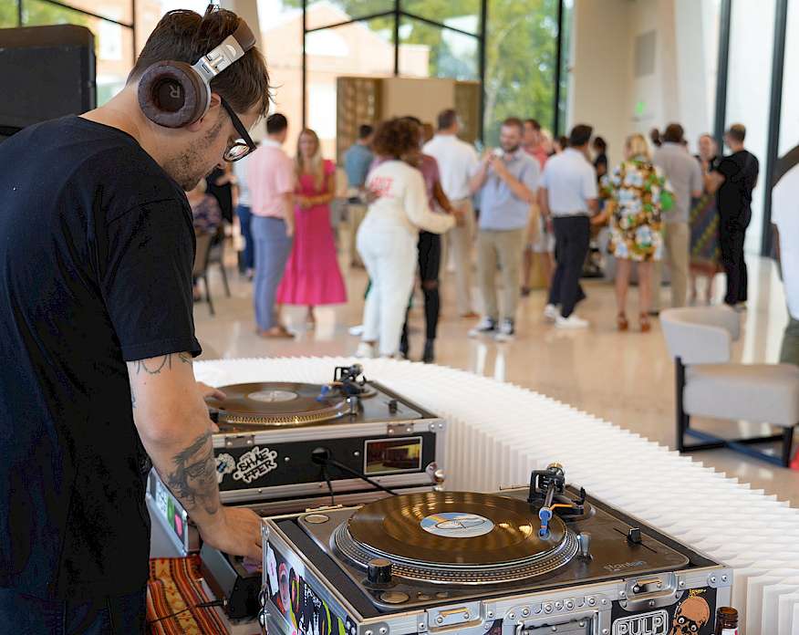 Photo of a DJ mixing records on a set of turntables with groups of people mingling and dancing at a party in the background.