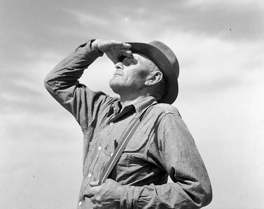 Black and white photograph of a man wearing a cowboy hat looking out in the distance with one hand up shielding his eyes.
