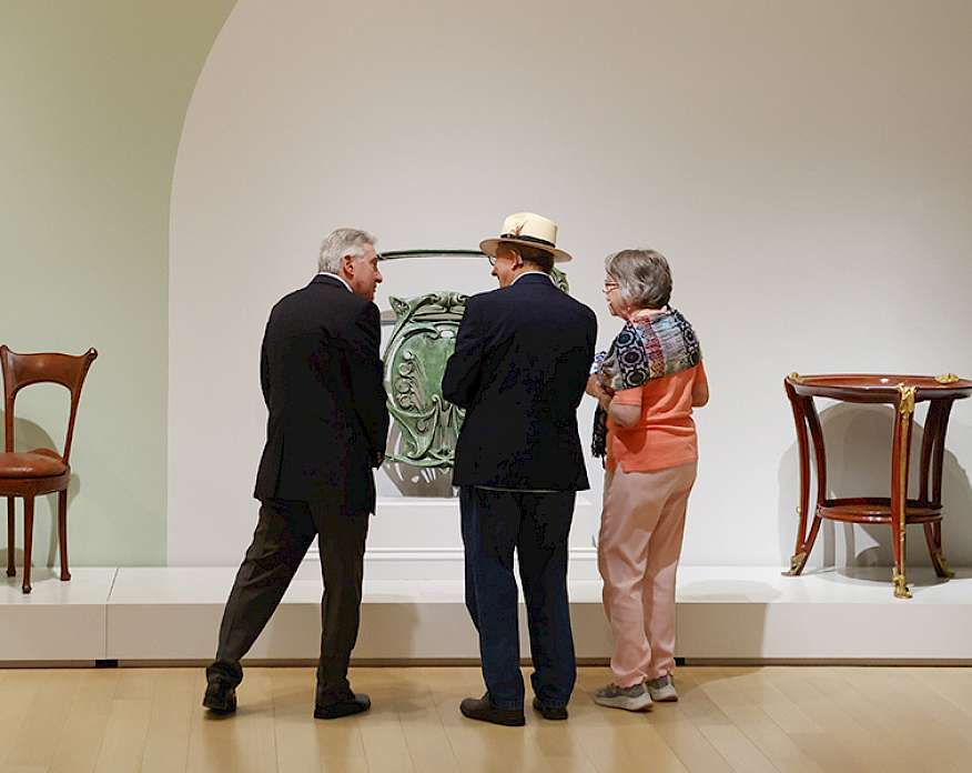 Photo of two men and one woman looking at Art Nouveau furniture in an art gallery.