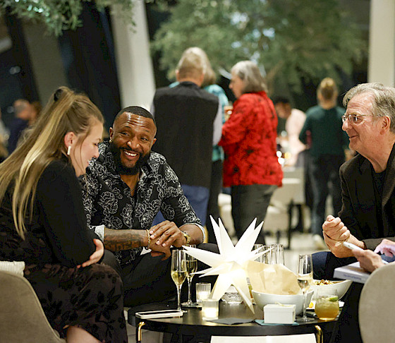 Photo of two men and two women sitting an a table drinking champagne with other people in the background.