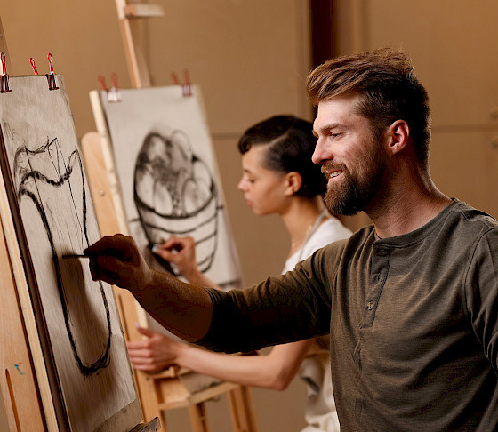 Photo of a man drawing a vase using charcoals on a sheet of paper at an art easel with a woman in the background seated drawing at another easel.