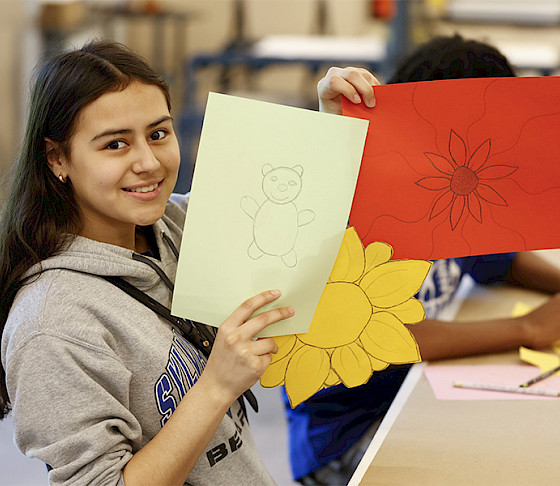 Photo of a young girl smiling and holding up drawings of flowers on colored construction paper.