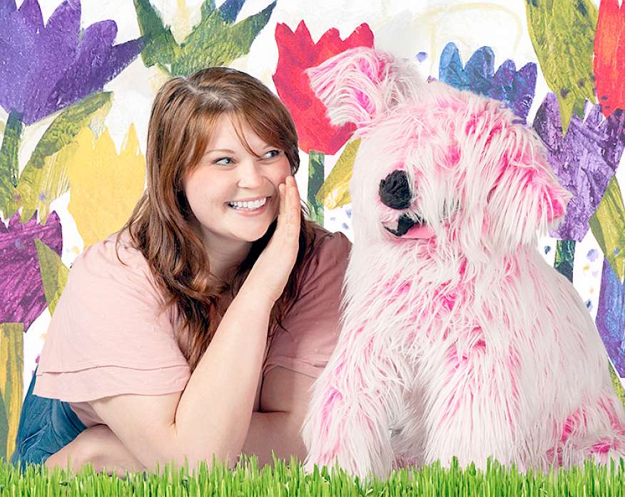 Photo of a woman whispering into the cocked ear of a pink puppet dog in front of a flowery background.