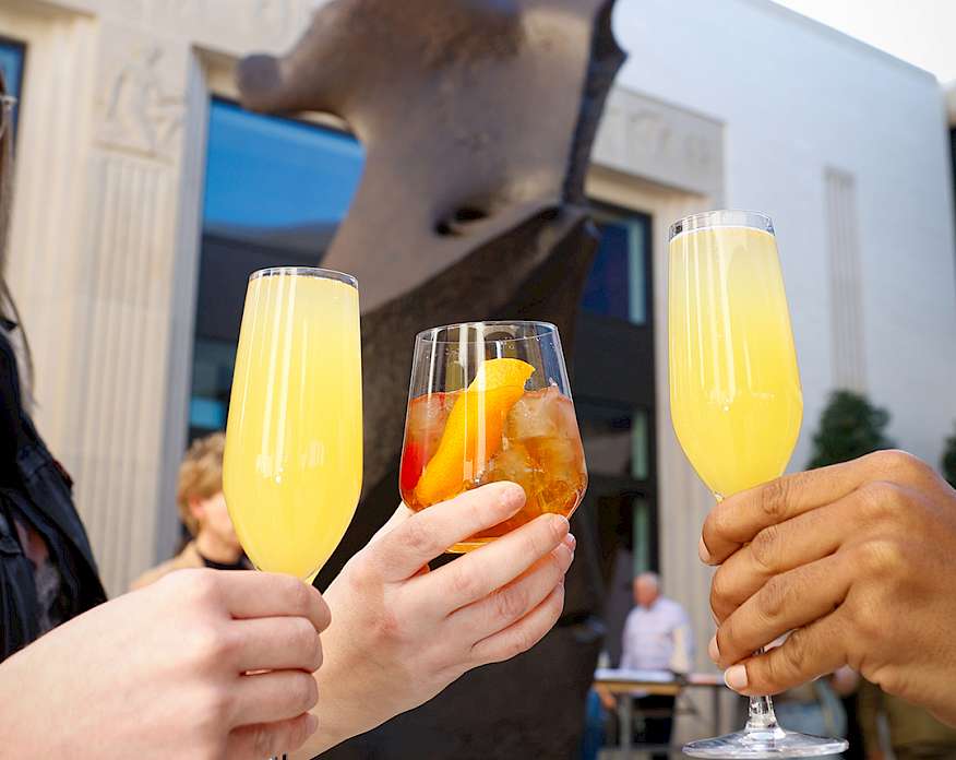Photo of three hands holding colorful cocktails next to a bronze sculpture.