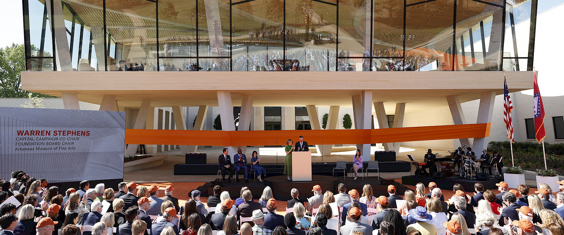 Photo of Harriet and Warren Stephens at a podium on a stage in front of a large orange ribbon wrapped around the courtyard entrance to AMFA on opening day.