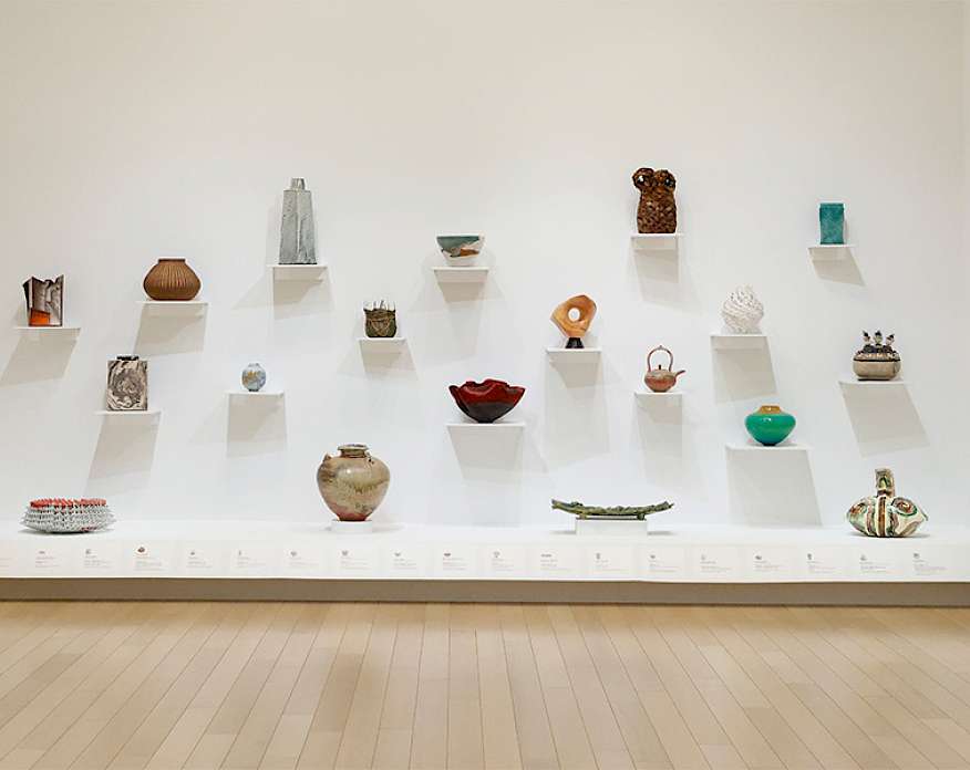 Photo of a white wall in an art gallery at AMFA displaying small vases and sculptures on white shelves.