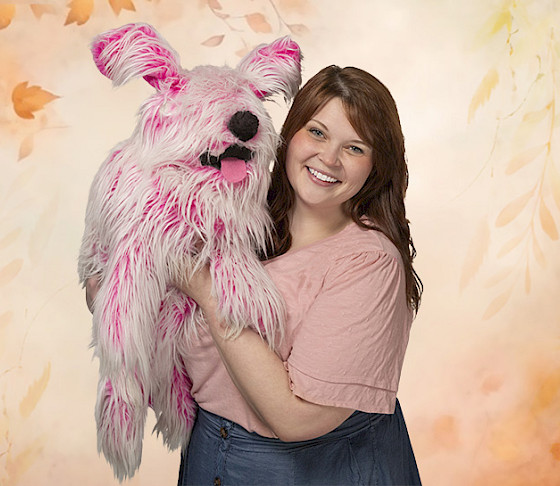 Photo of a woman holding a pink puppet dog in front of a leafy background.
