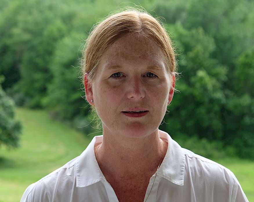 Photo of Anne Lindberg wearing a white shirt with her red hair pulled back standing in front a green meadow with trees.