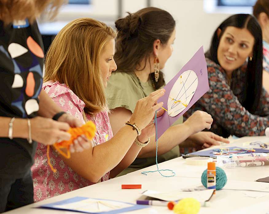 Photo of several white women sitting at a white table doing an art activity with paper, yarn, and glue sticks.