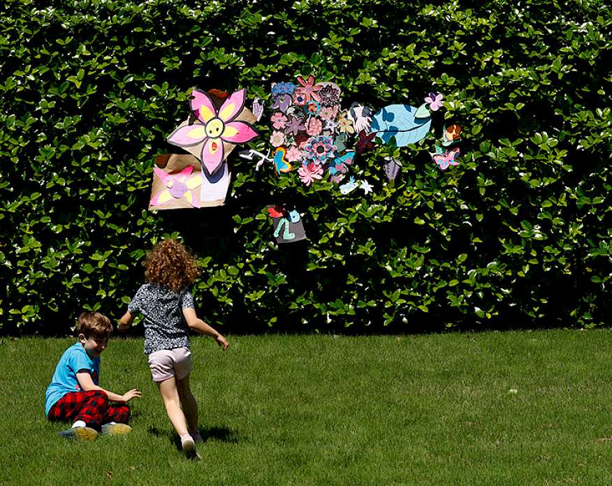 Photo of a large hedge decorated with handmade paper flowers. A young boy and young girl sit playing in front of it.