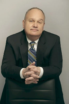 Photo of Brian Lang wearing a black suit, white shirt, and blue and yellow striped tie standing with his hand laced together on top of the back of a black leather chair.