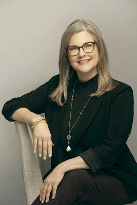 Photo of Catherine Bays wearing a black dress and long gold necklace sitting on a beige chair.