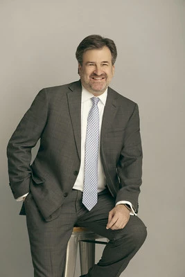 Photo of Laine Harper wearing a medium gray suit, white shirt, and light blue tie sitting on a metal stool.