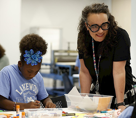 Photo of a teacher smiling and standing next to a table where several young children are doing art projects.