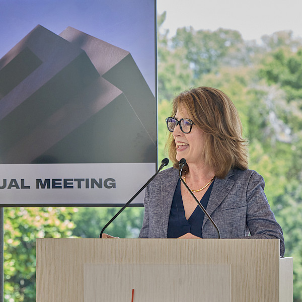 Photo of Dr. Victoria Ramirez speaking at a podium with a screen behind her displaying a photo of the Arkansas Museum of Fine Arts with the words 2025 AMFA Annual Meeting.