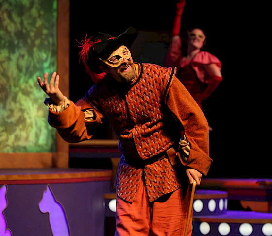Photo of an actor performing in a Children's Theatre show at AMFA. He wears a black hat with a red feather, an orange Shakespearian-era outfit, and a gold mask while holding a light wooden cane.