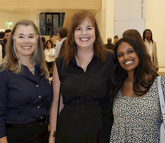 Photo of three women smiling in the Atrium of AMFA surrounded by other people during an event.